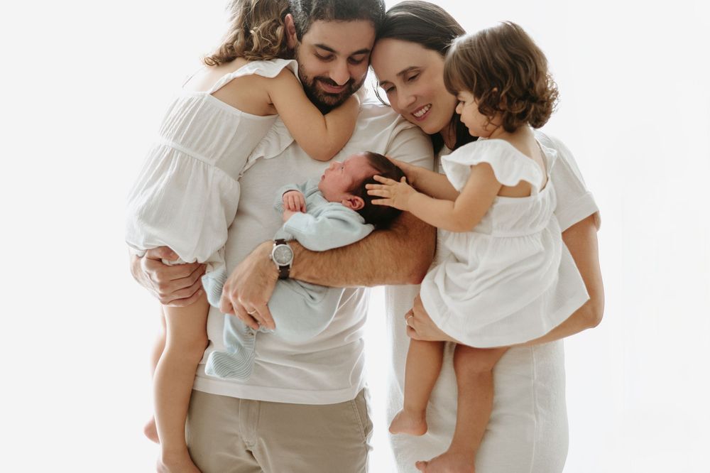 Happy family in white outfits cuddles newborn baby together against a white background.