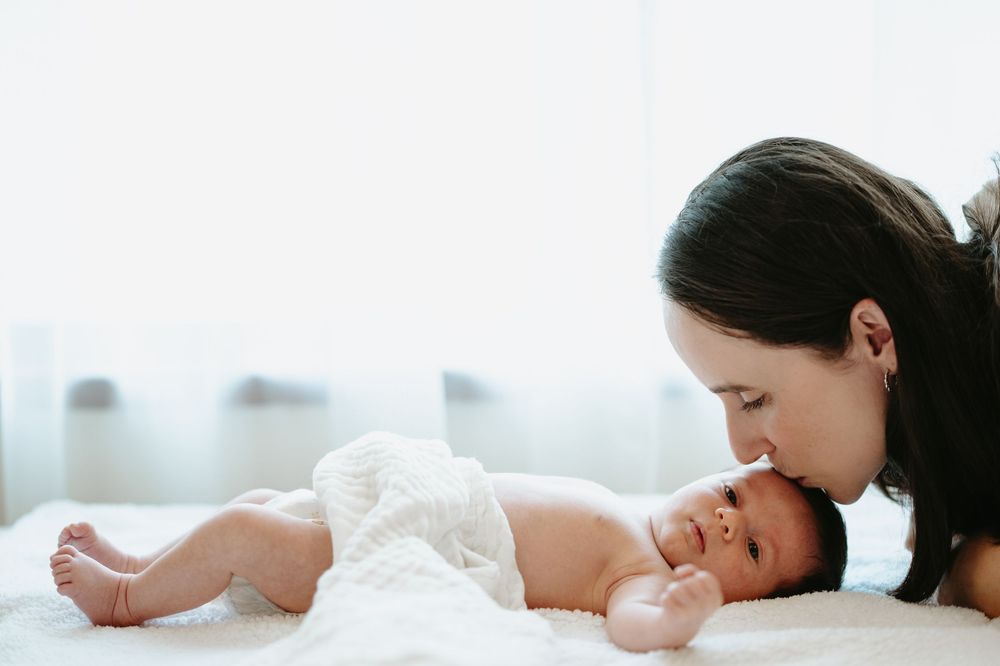 A mother tenderly kisses her newborn baby lying on a white blanket in a soft, bright room.