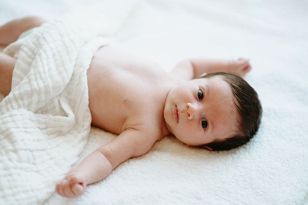 A peaceful newborn baby with dark hair lying on a soft white blanket, gazing curiously at the camera.