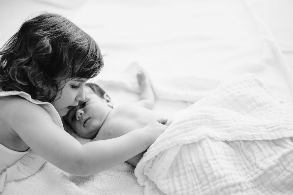 Black and white photo of a young child tenderly holding a sleeping newborn baby wrapped in a soft white blanket.