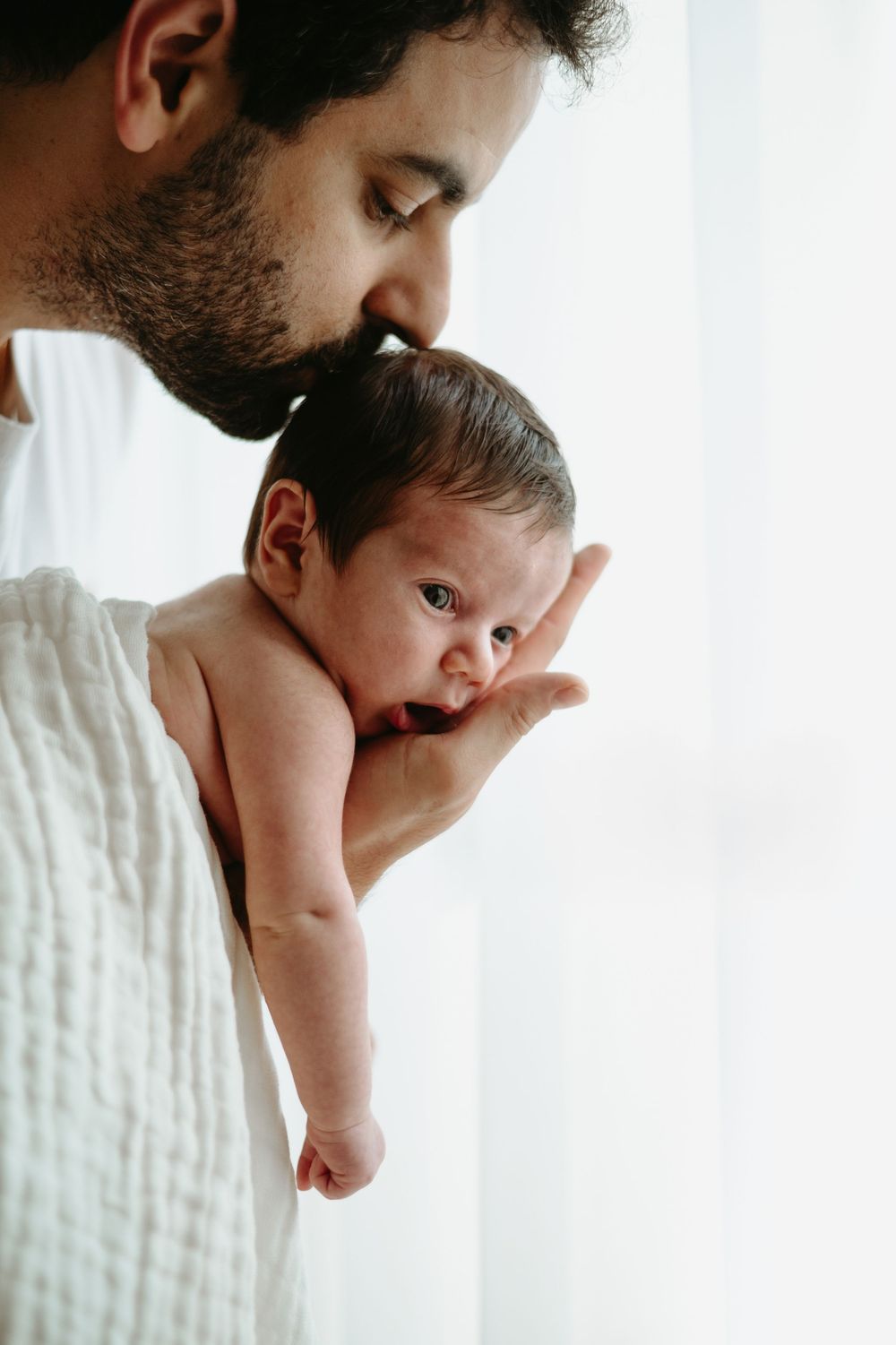 Bearded father tenderly kissing his newborn baby while gently cradling the infant in his hands.