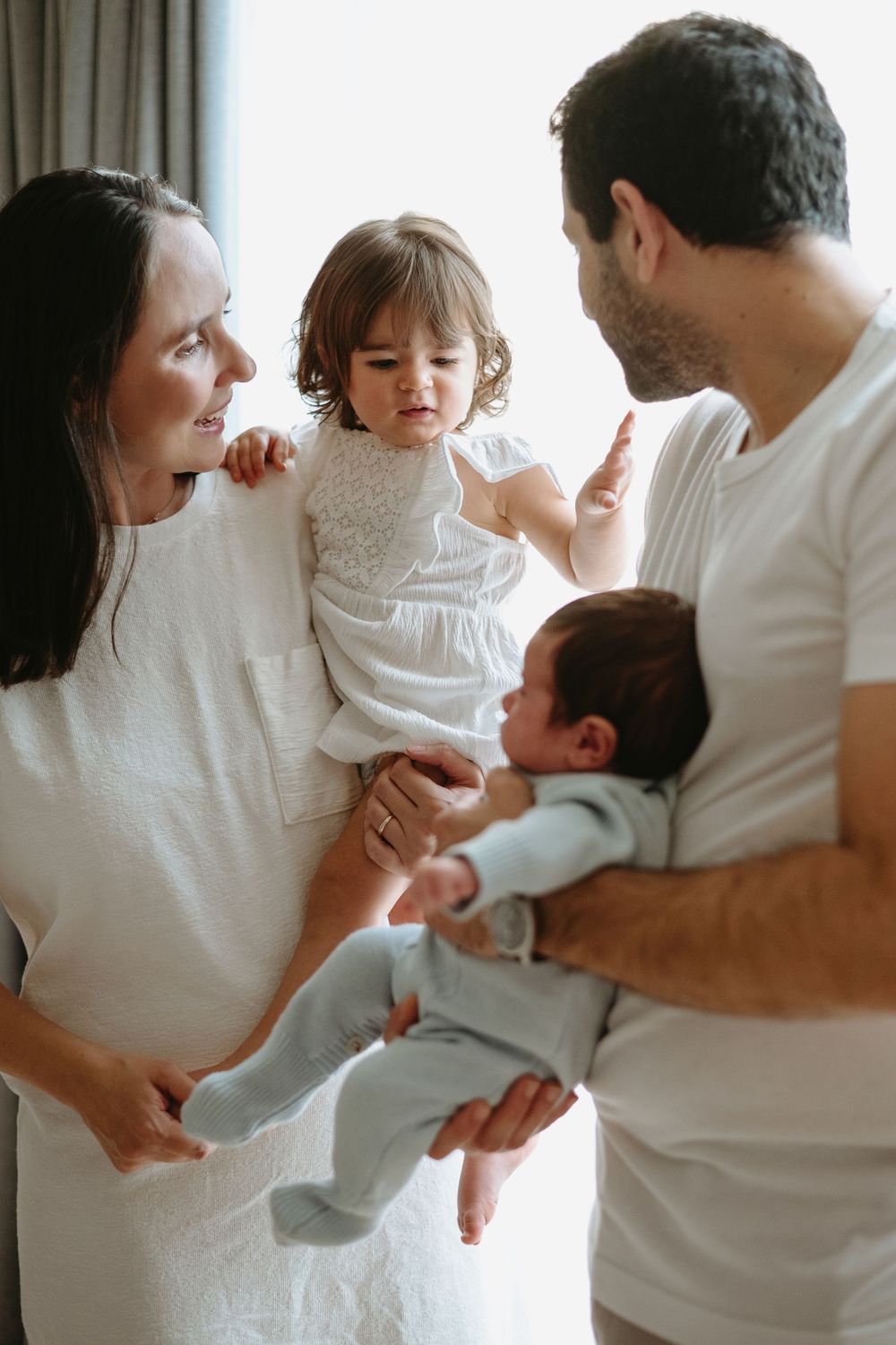 A young family with a toddler in white dress and newborn baby sharing a tender moment indoors.