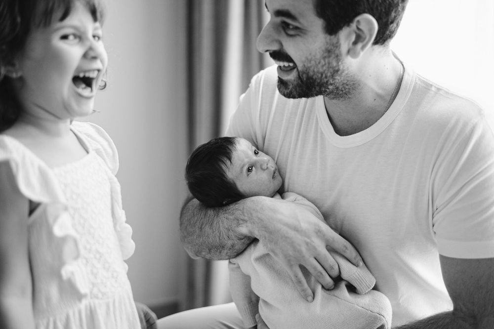 Black and white photo of a father holding a newborn baby while smiling at his excited toddler daughter.