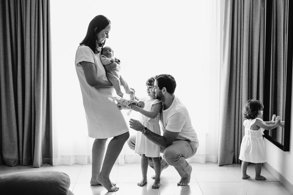 Black and white family photo of parents with newborn baby and toddler daughter in bright room.