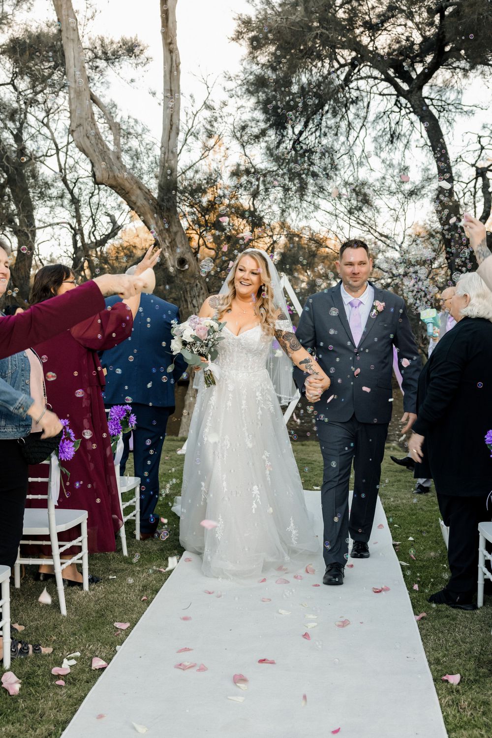 Bride and groom walk down outdoor aisle as guests toss rose petals at their wedding ceremony.