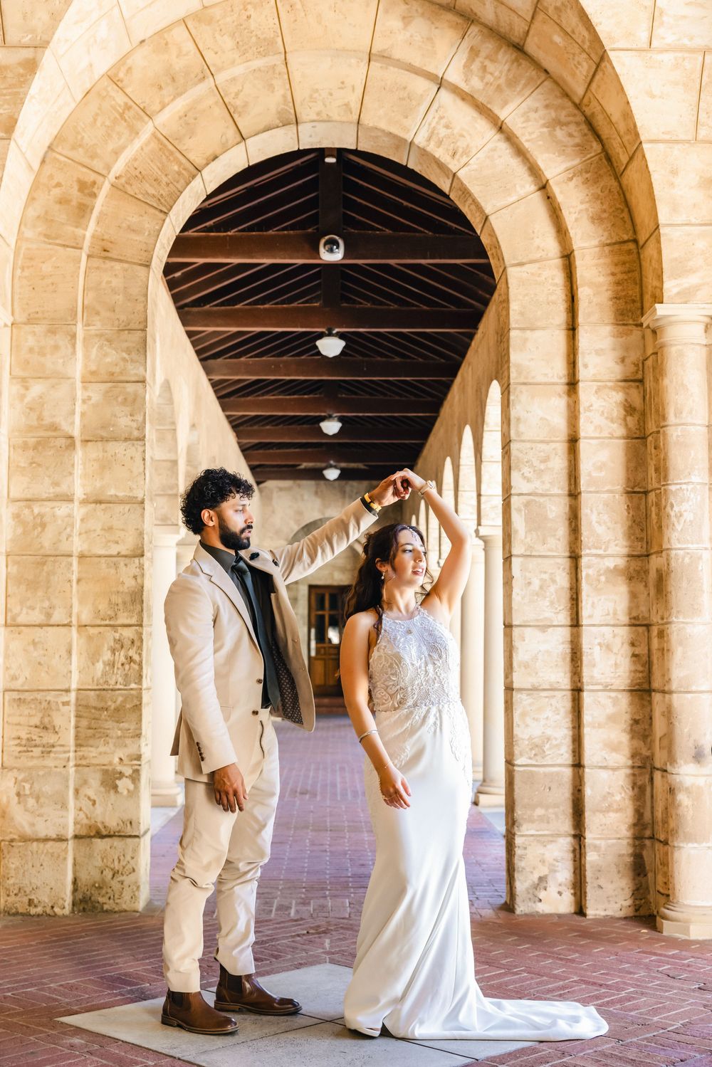 Bride and groom dancing under stone arches in a elegant outdoor corridor, bride in white gown, groom in beige suit.