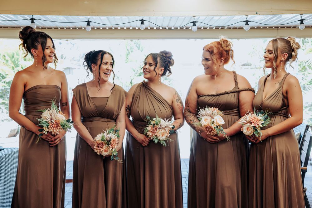 Bridesmaids in matching brown dresses holding floral bouquets at outdoor wedding.