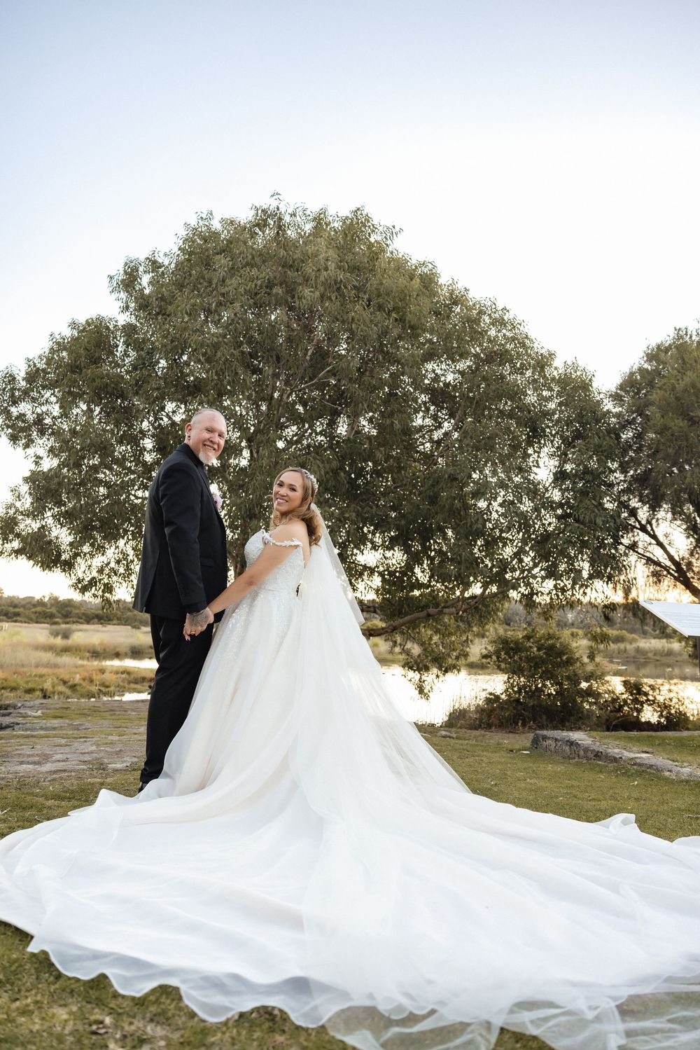 Bride in flowing white gown and groom in dark suit pose outdoors near a tree and lake on their wedding day.
