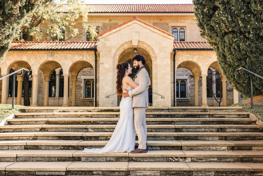 Couple in wedding attire embracing on stone steps before a grand arched building with lush trees.