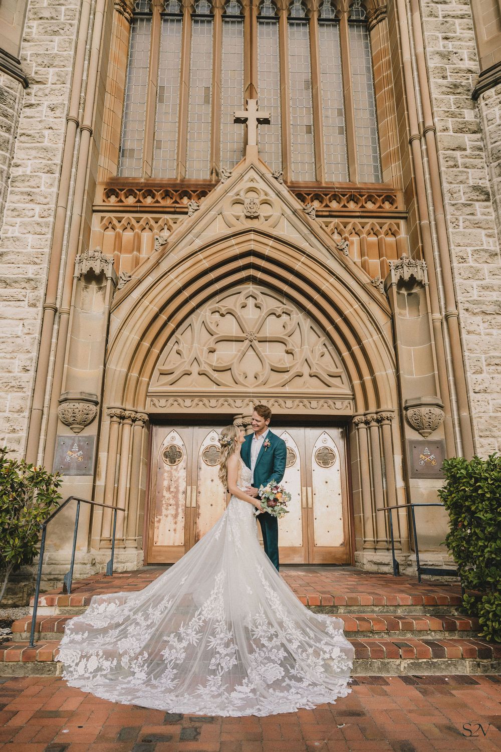 Bride and groom pose before ornate Gothic church entrance, her lace cathedral train cascading down brick steps.