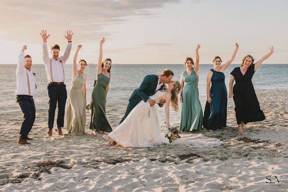 Wedding party celebrating on a beach, bridesmaids in green dresses, groomsmen in navy, couple kissing at sunset.