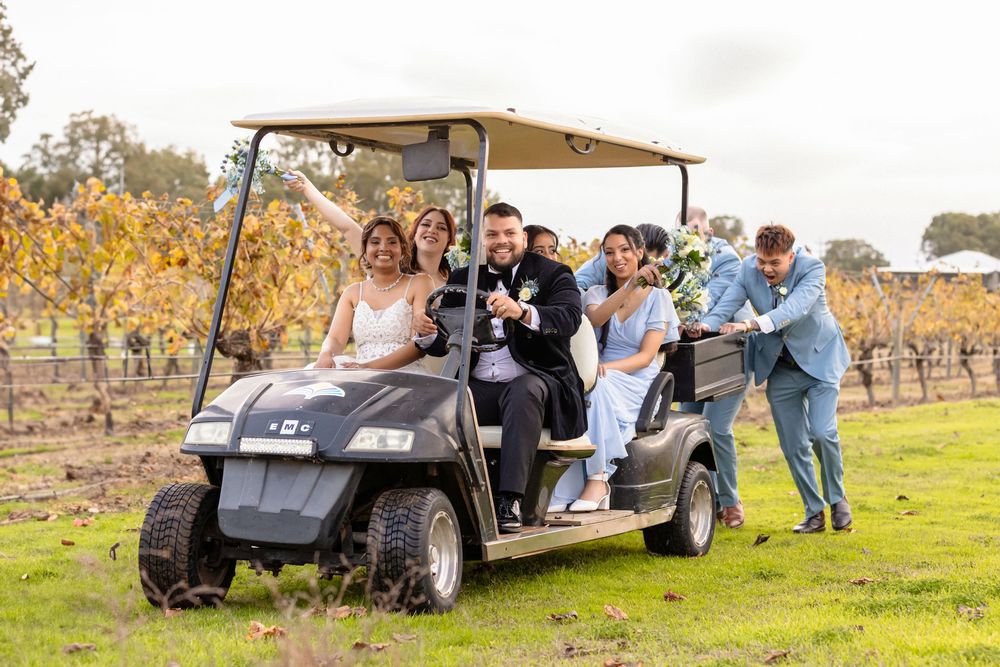 A group of wedding guests laugh and push a golf cart through scenic vineyard grounds on a fall day.