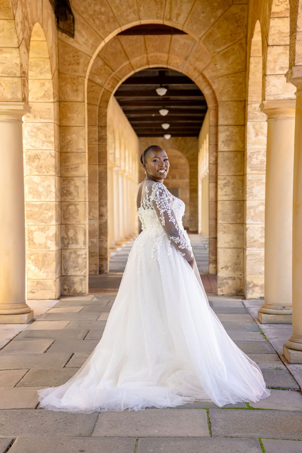 Elegant bride in flowing white wedding gown poses in a sunlit stone colonnade at Stanford University.