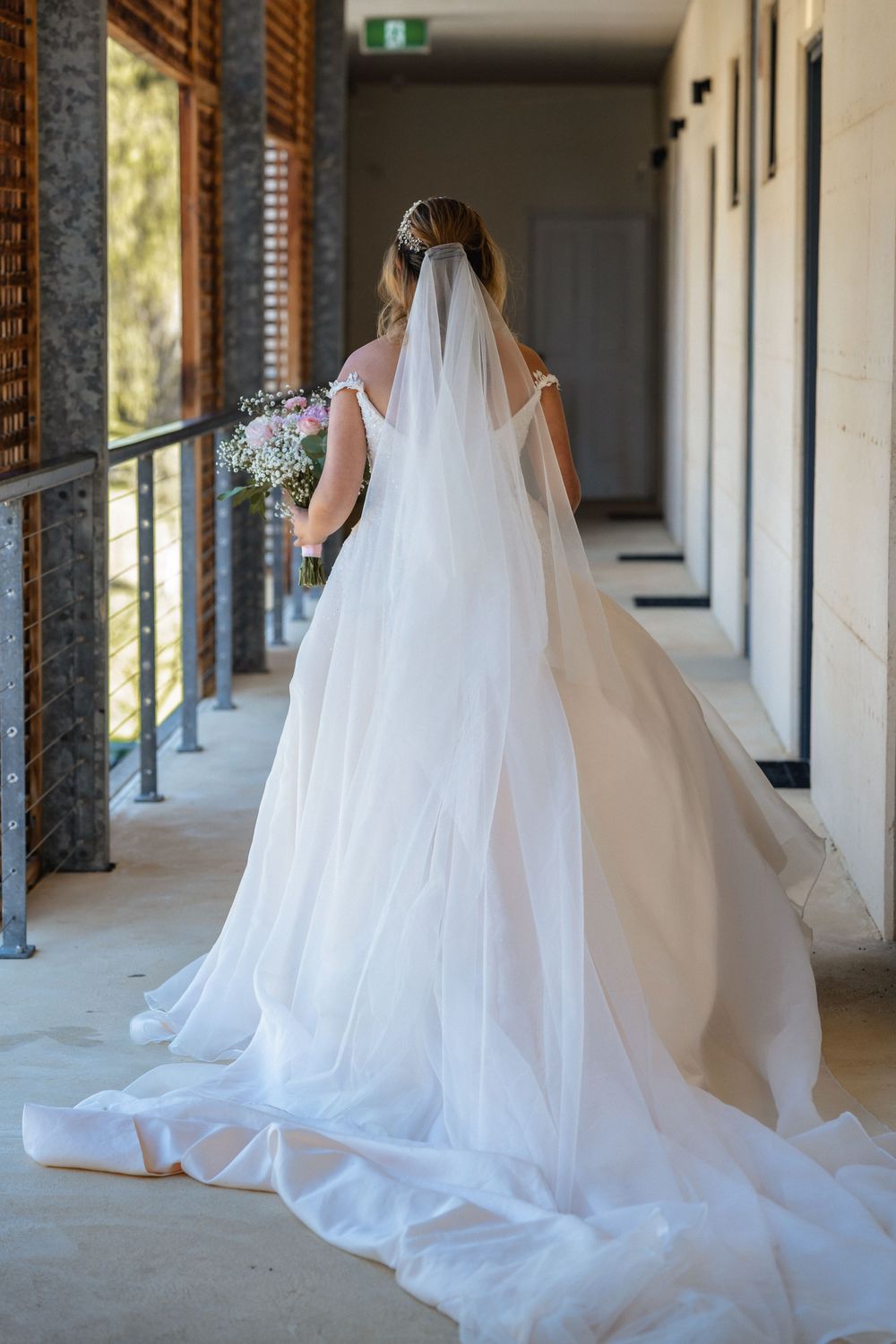Bride in white wedding gown with long veil and train walks down outdoor corridor holding bouquet.