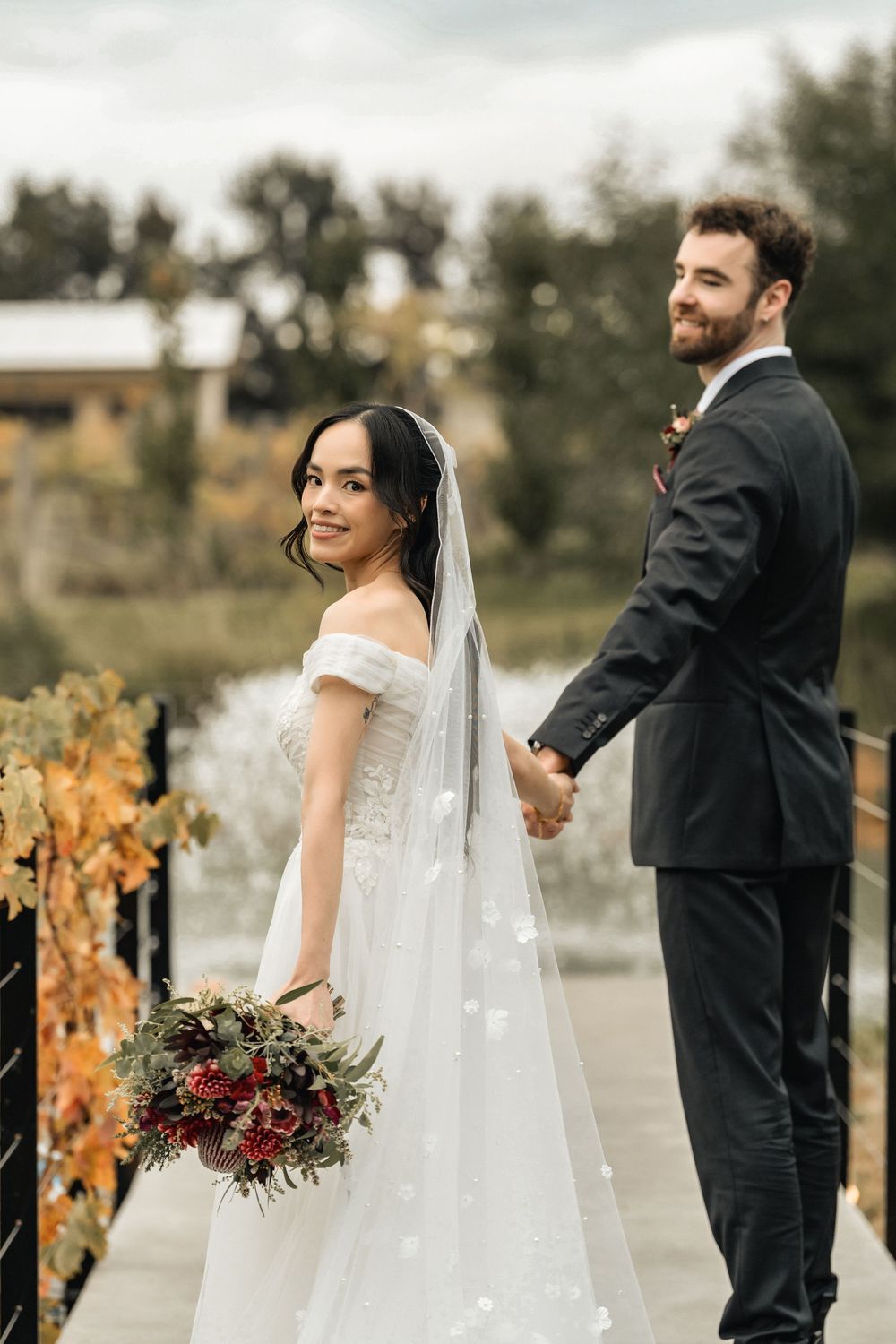 Bride and groom holding hands on a bridge, bride in off-shoulder gown with veil, holding autumn bouquet.