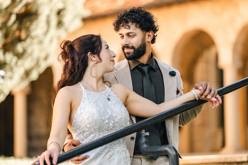 A bride and groom share a romantic moment leaning on a railing outdoors at a sunny wedding venue.