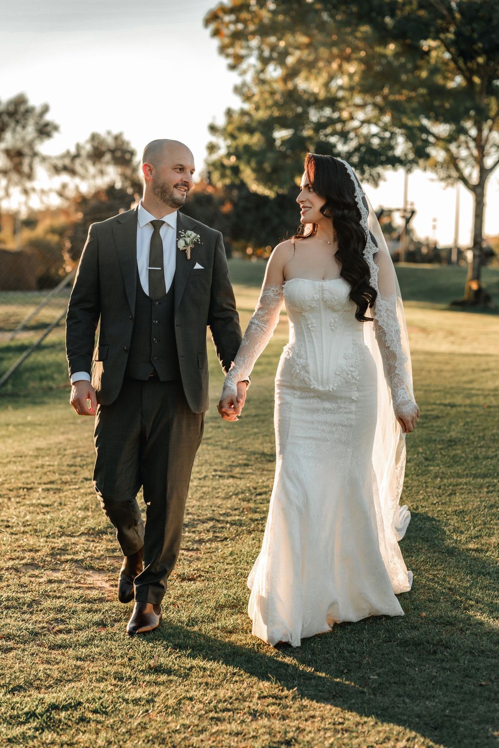Smiling bride and groom walk hand in hand across a sunlit green lawn, bride in a fitted lace wedding dress.