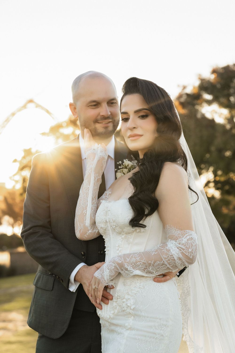 A bride and groom pose outdoors at golden hour, she wears a lace gown and veil, he wears a dark suit.