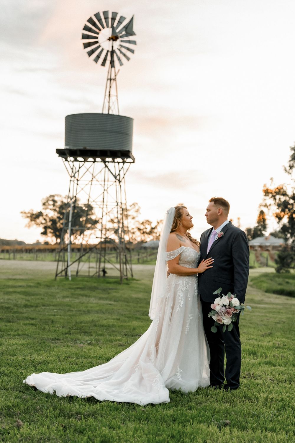 Bride and groom embracing at sunset on green grass with a rustic windmill water tower in the background.