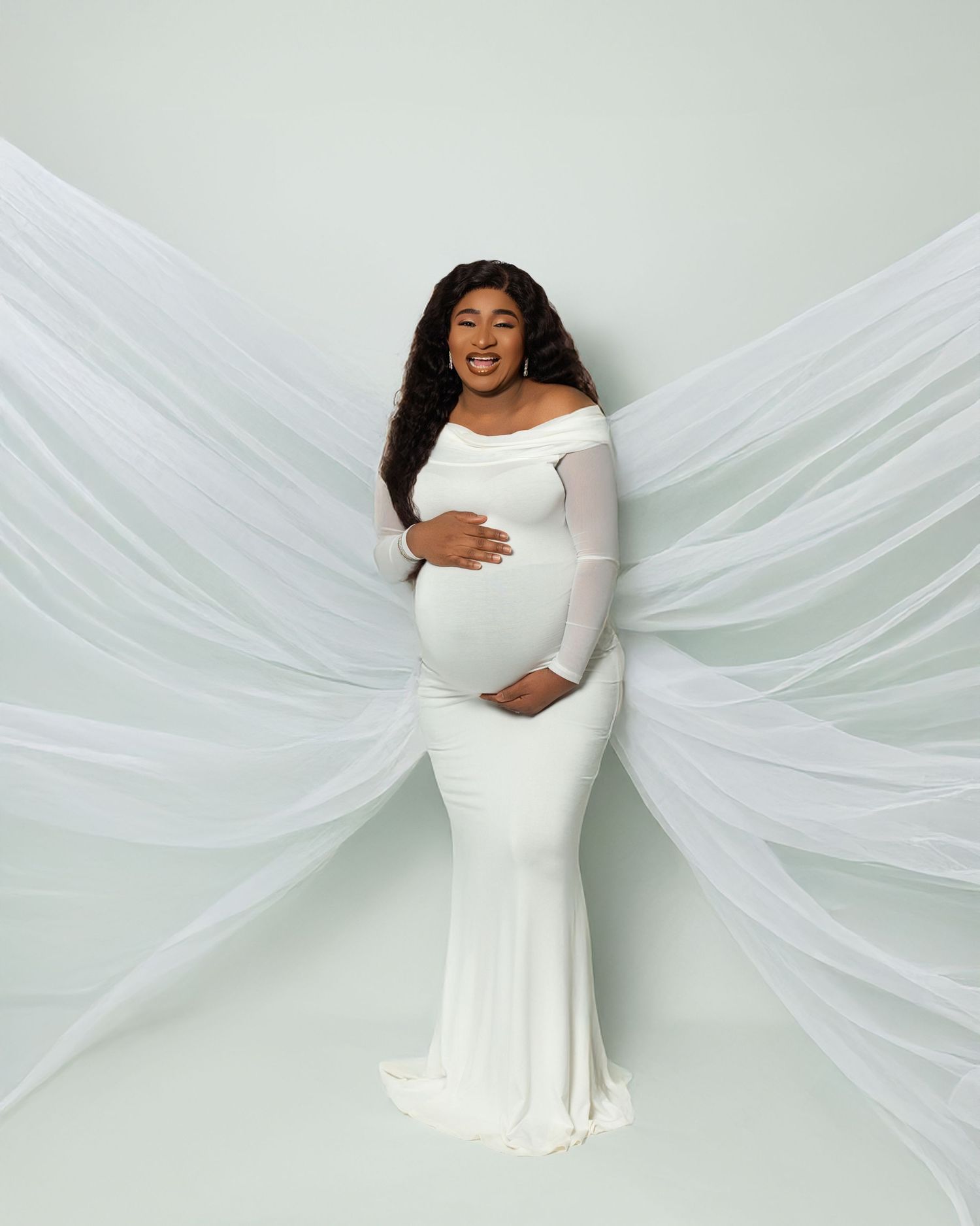 Ethereal maternity portrait featuring an elegant white off-shoulder gown with flowing tulle fabric against a light backdrop.