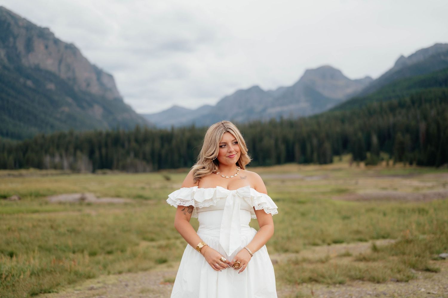 Smiling blonde woman in white off-shoulder dress posing in mountain meadow with dramatic peaks behind.
