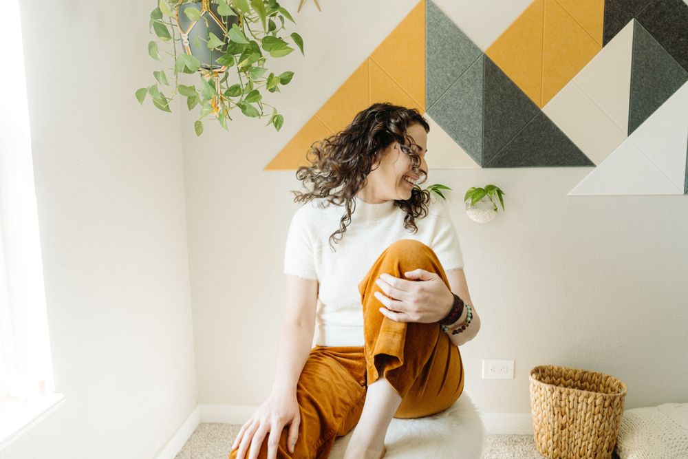 Woman in white shirt and brown pants sitting by geometric wall pattern and plants.