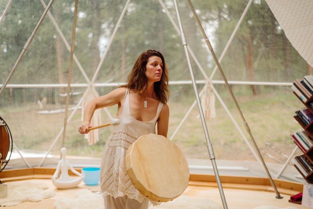 Woman in flowing dress holding drum inside geodesic dome structure.