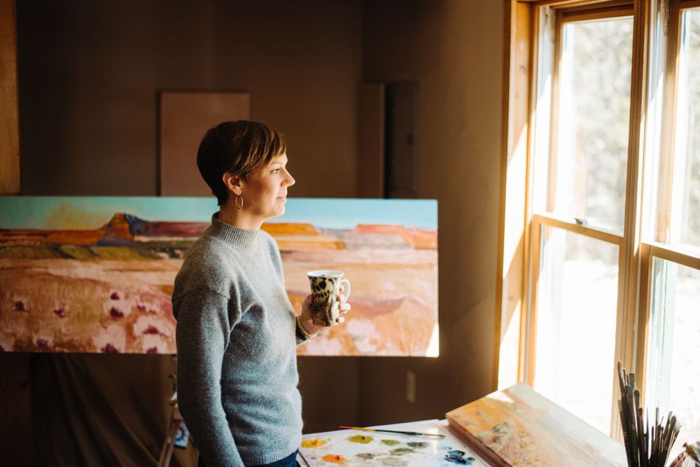 Artist sipping coffee while looking out studio window with painting in background.
