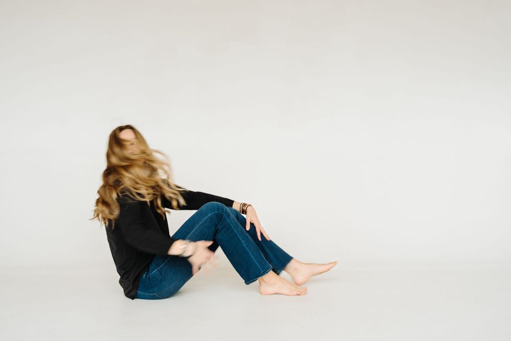 Person in black sweater and jeans sitting on floor against white background with flowing blonde hair.