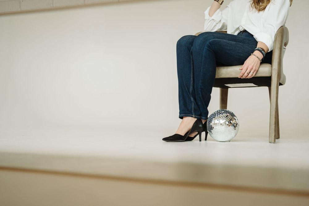 Person sitting in chair wearing blue jeans and black high heels with silver metallic purse on floor.