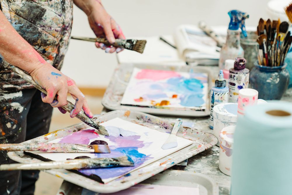 Artist using watercolors and brushes to create colorful abstract paintings on a messy workspace.