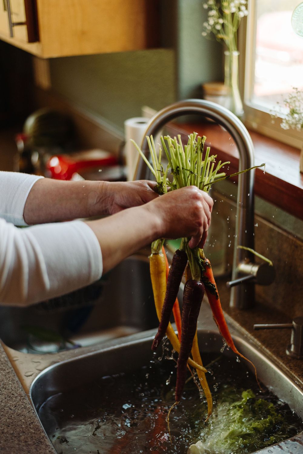 Hands washing fresh colorful carrots in kitchen sink with running water.