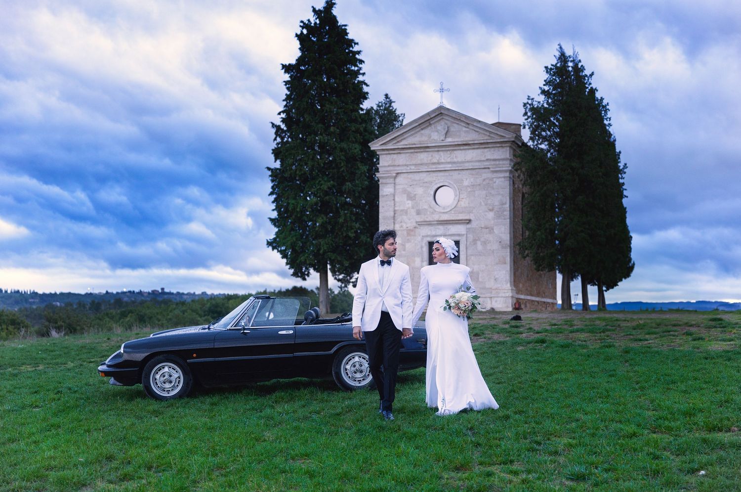 Romantic outdoor scene with couple embracing near chapel in Tuscan countryside.