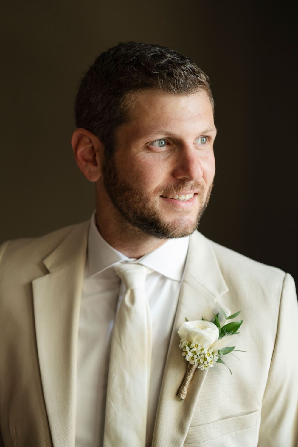 A groom in a cream suit and white boutonniere smiles warmly in this wedding portrait with soft lighting.