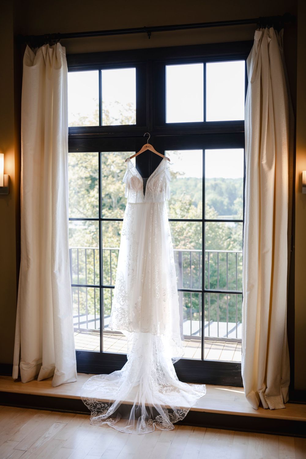 White lace wedding dress with long train hanging in window with sheer curtains and natural light streaming in.
