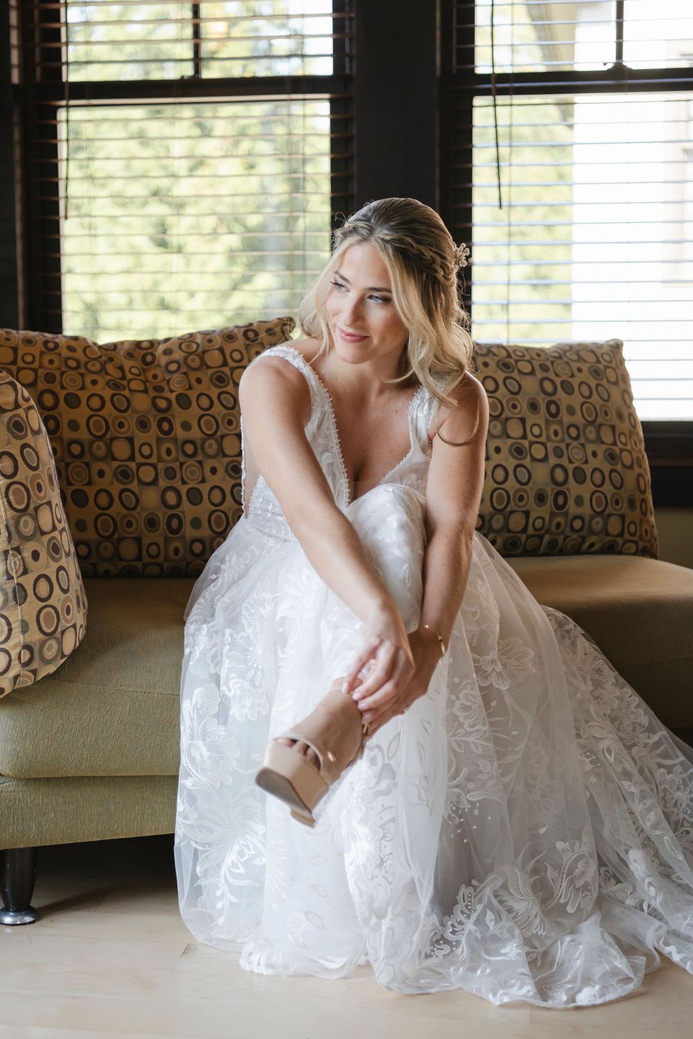 A bride in a flowing white lace wedding gown sits on a patterned couch by a window, adjusting their shoe.