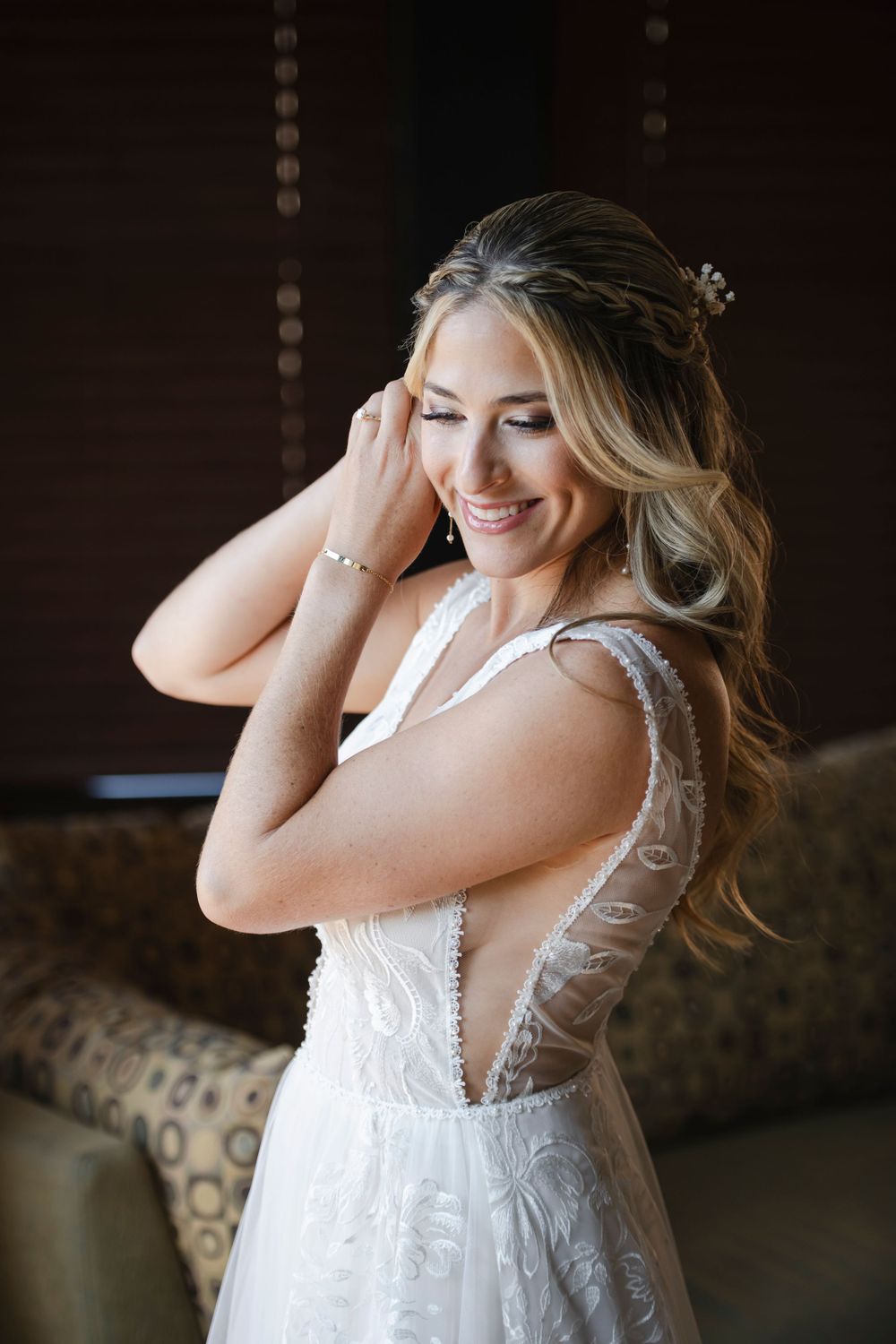 Bride wearing delicate lace wedding dress smiles while putting in her earrings in natural light setting.