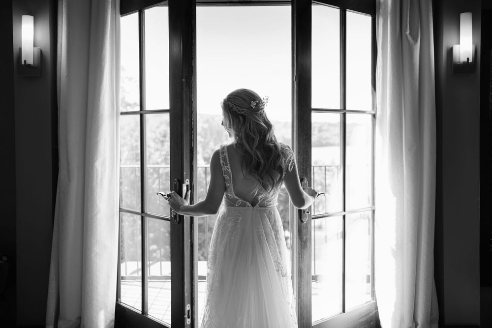 A bride in a flowing white dress stands silhouetted against French doors and natural light in black and white.