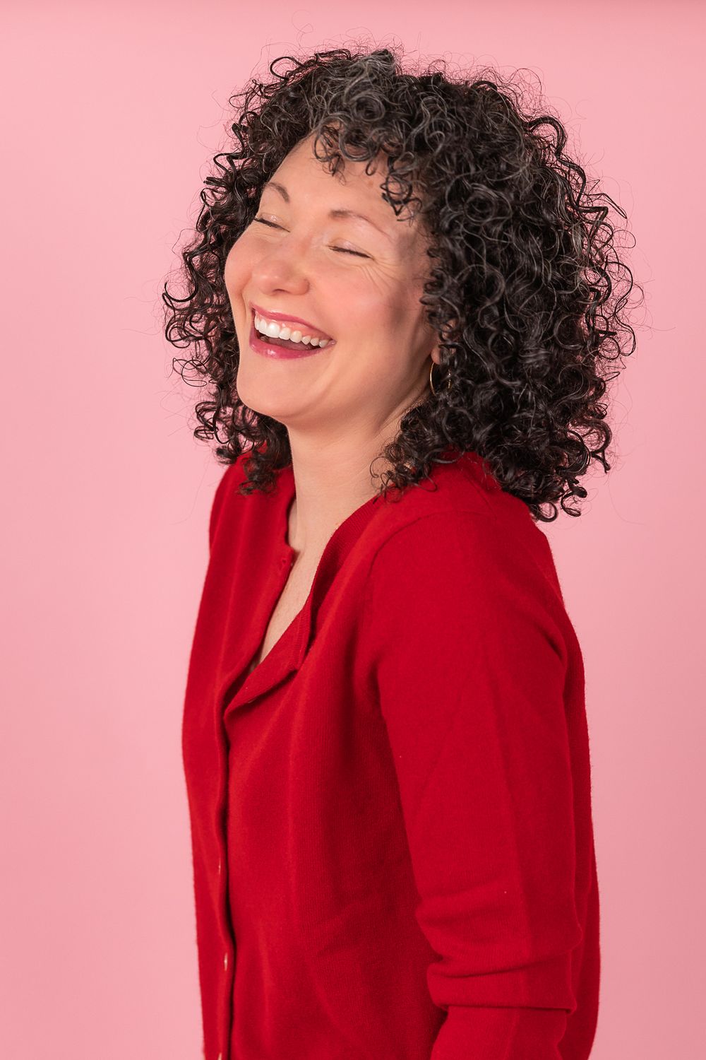 A person with curly dark hair wearing a red blazer laughs joyfully against a pink background.