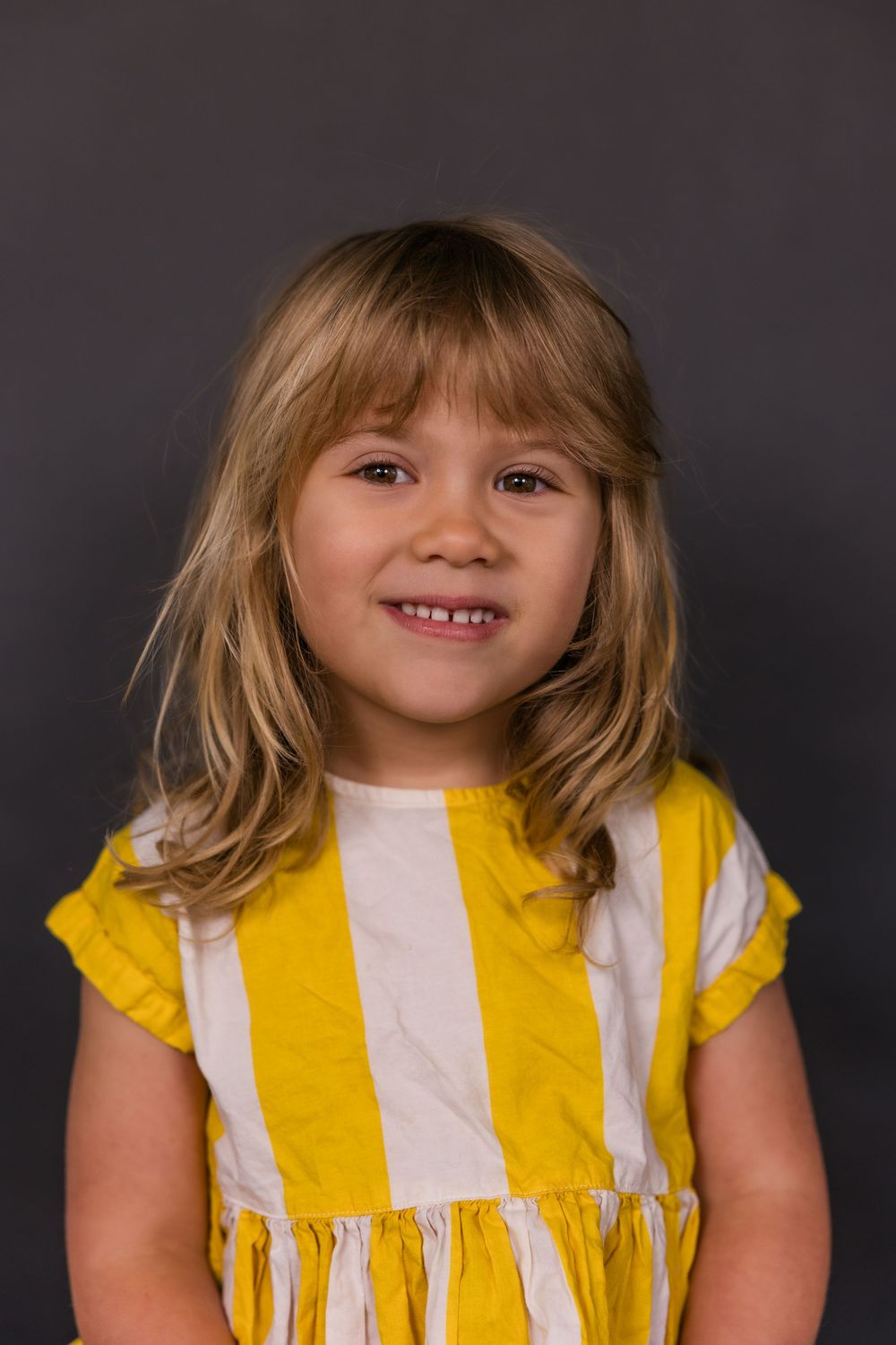Child wearing yellow and white striped dress smiles warmly against dark background in studio portrait.
