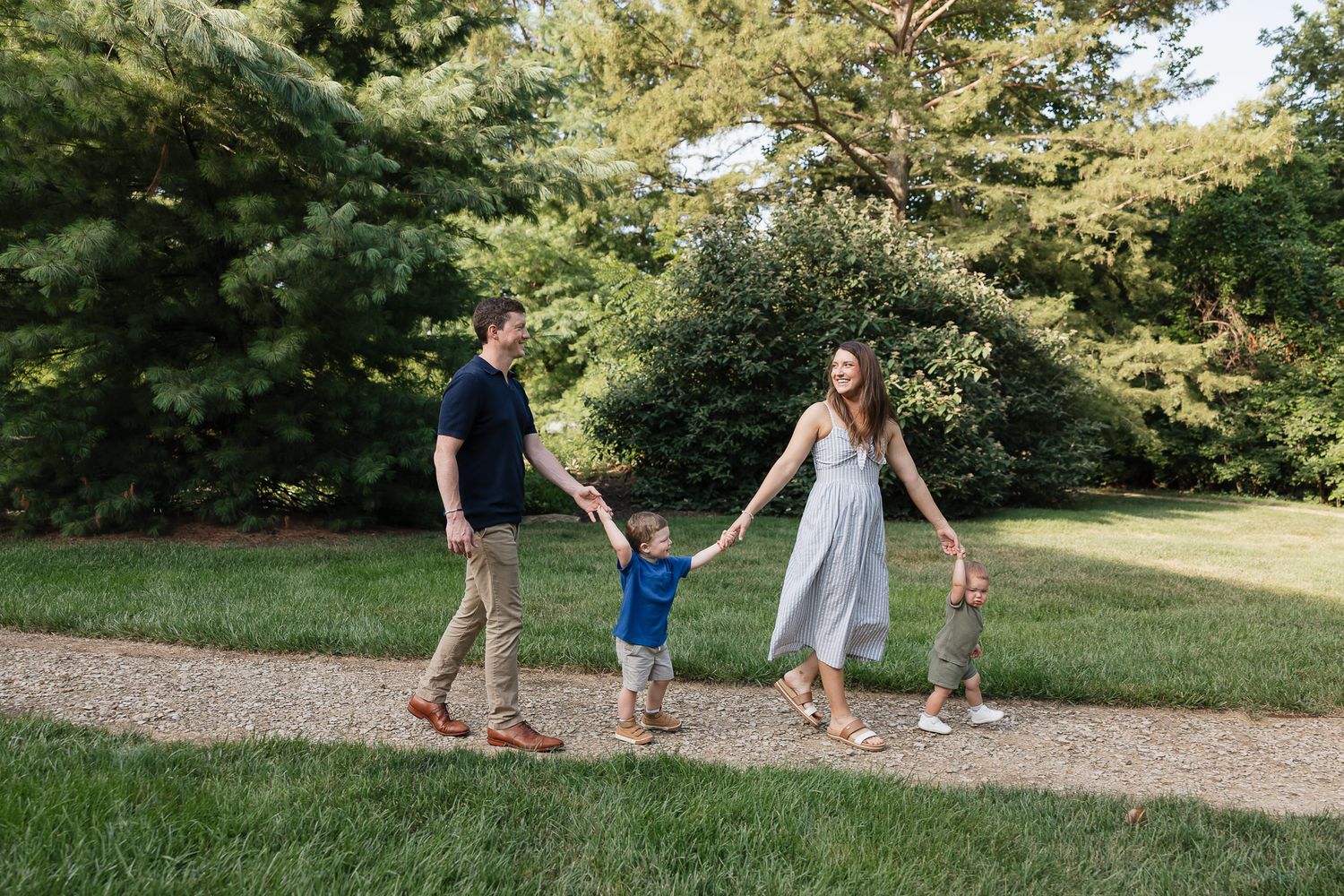 Photographer overland park ks takes photo of family of 4 walking on gravel path holding hands with trees behind them at antioch park