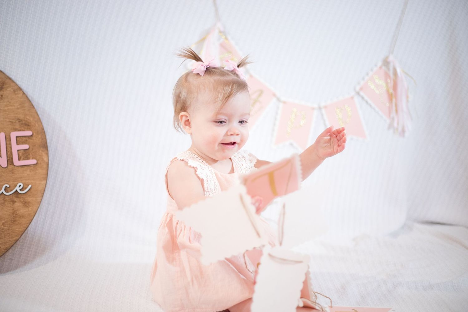 baby in pink bow and dress reaching toward birthday decorations in a white studio.