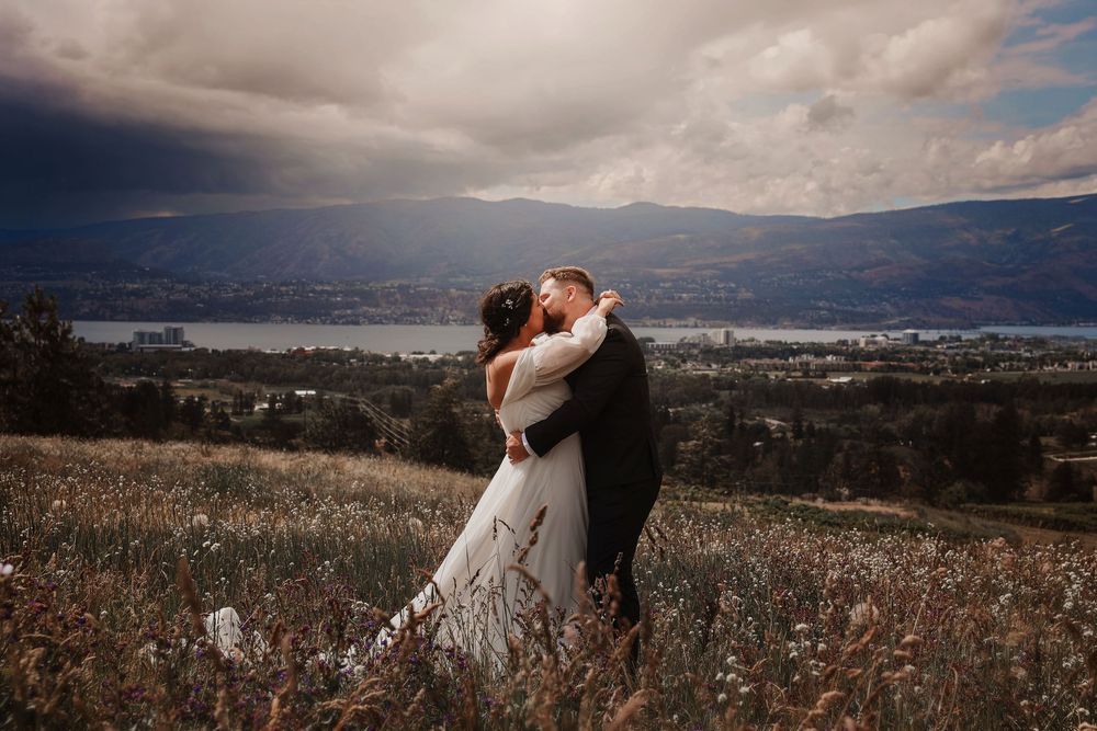 Couple embracing on a hillside meadow with dramatic stormy skies and a scenic lake valley backdrop on their wedding day.