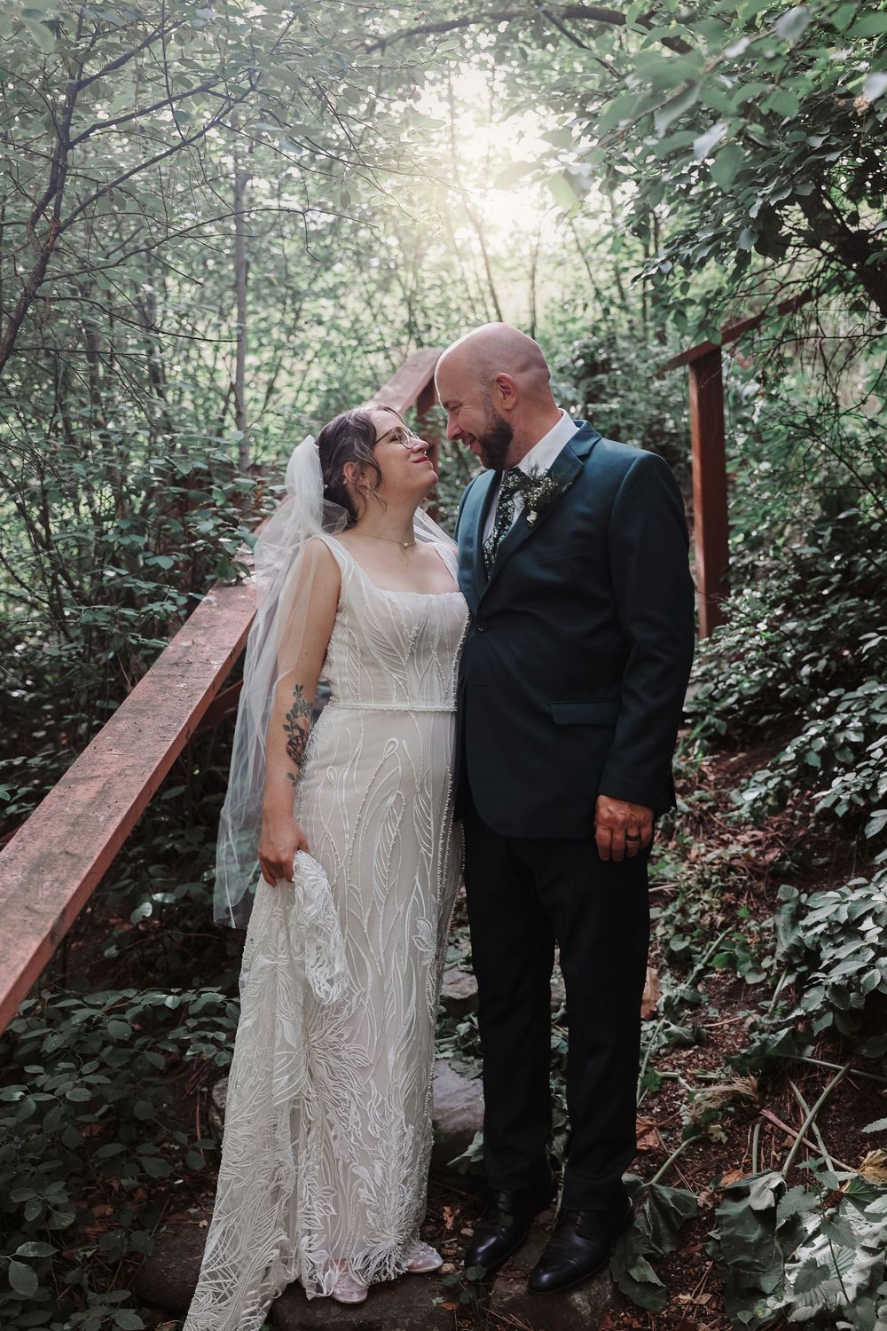 Bride and groom share a romantic moment on a wooden bridge surrounded by lush green forest on their wedding day.