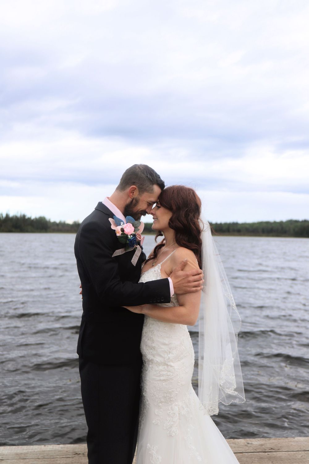 A romantic wedding couple shares a tender moment on a wooden dock by a lake.