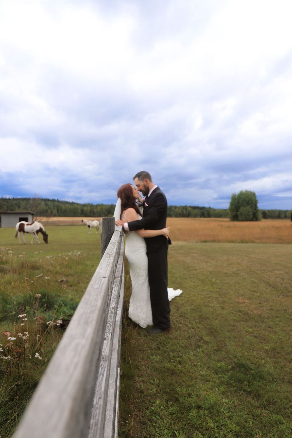 A couple shares a romantic moment in their wedding attire on a scenic rural field with cloudy skies.
