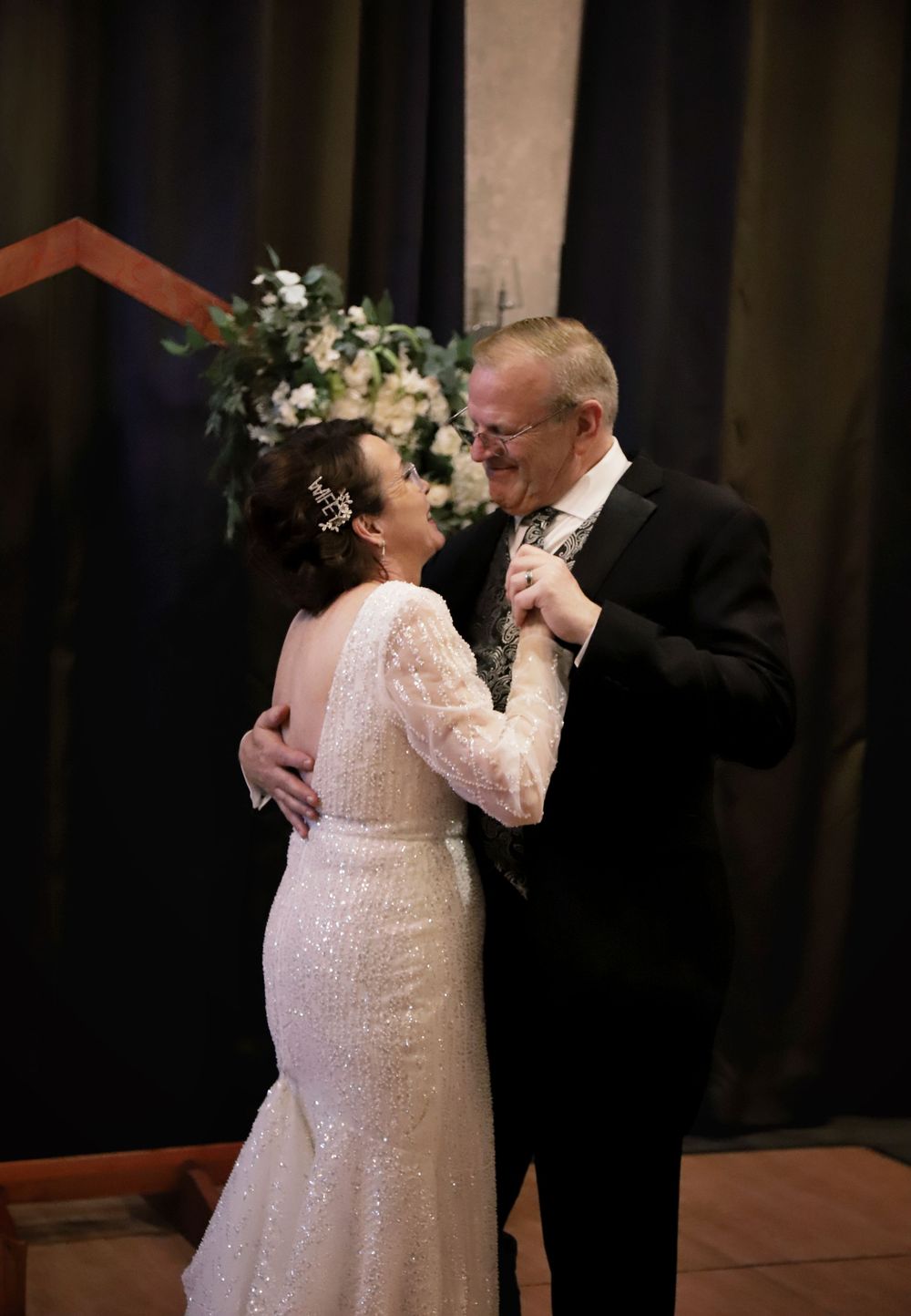 Couple shares intimate dance moment in elegant formal attire against dark backdrop.