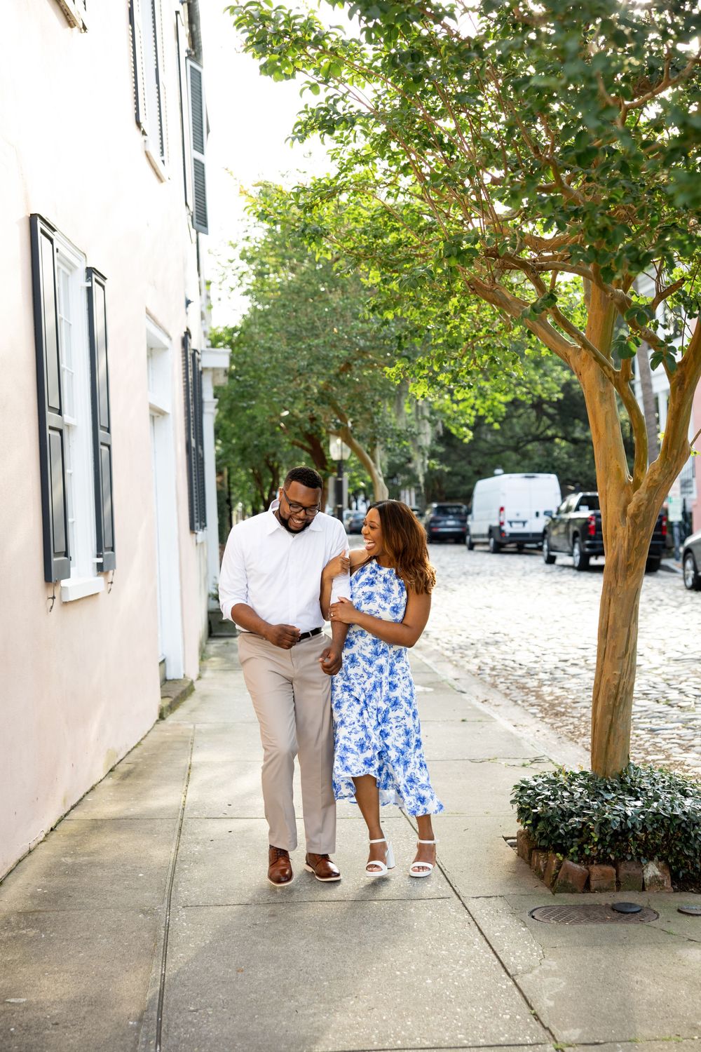 A couple in coordinated white and blue outfits walks down a tree-lined urban sidewalk.