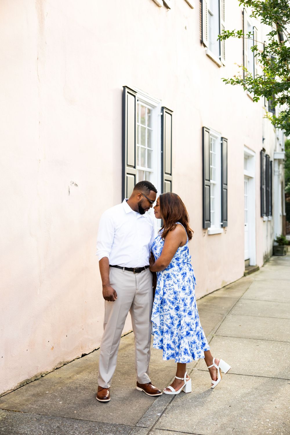 A couple in blue and white outfits walk together along a white stucco wall on a sunny day.