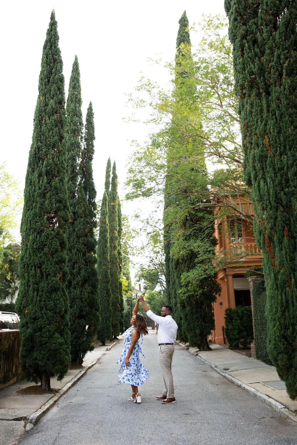 Two people stroll together on a street flanked by tall cypress trees and a terracotta-colored building.
